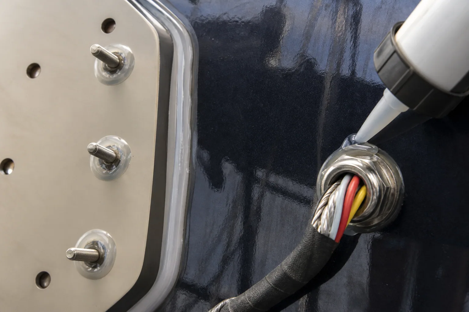 Marine sealant applied around stainless yacht letter studs and waterproof cable gland on dark gelcoat.